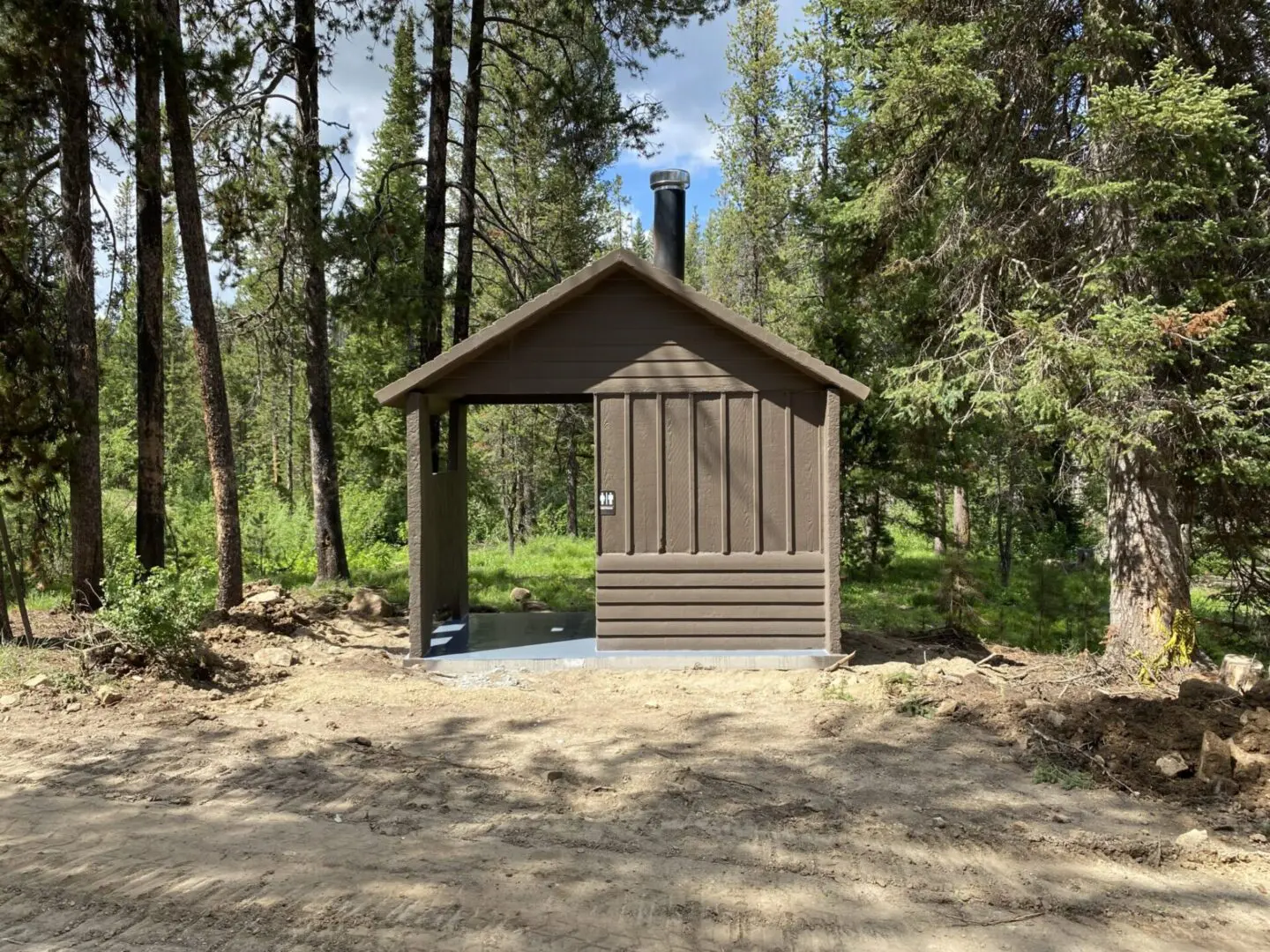 A small wooden outhouse is located in a forested area, surrounded by trees and a dirt path in the foreground.