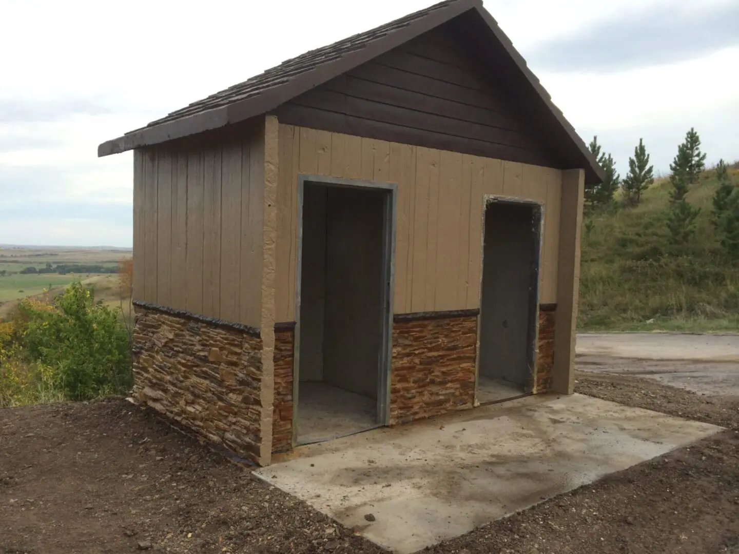 A small, simple wooden and stone building with two open doorways set on a concrete base, positioned on a grassy hillside with trees and a distant landscape in the background.