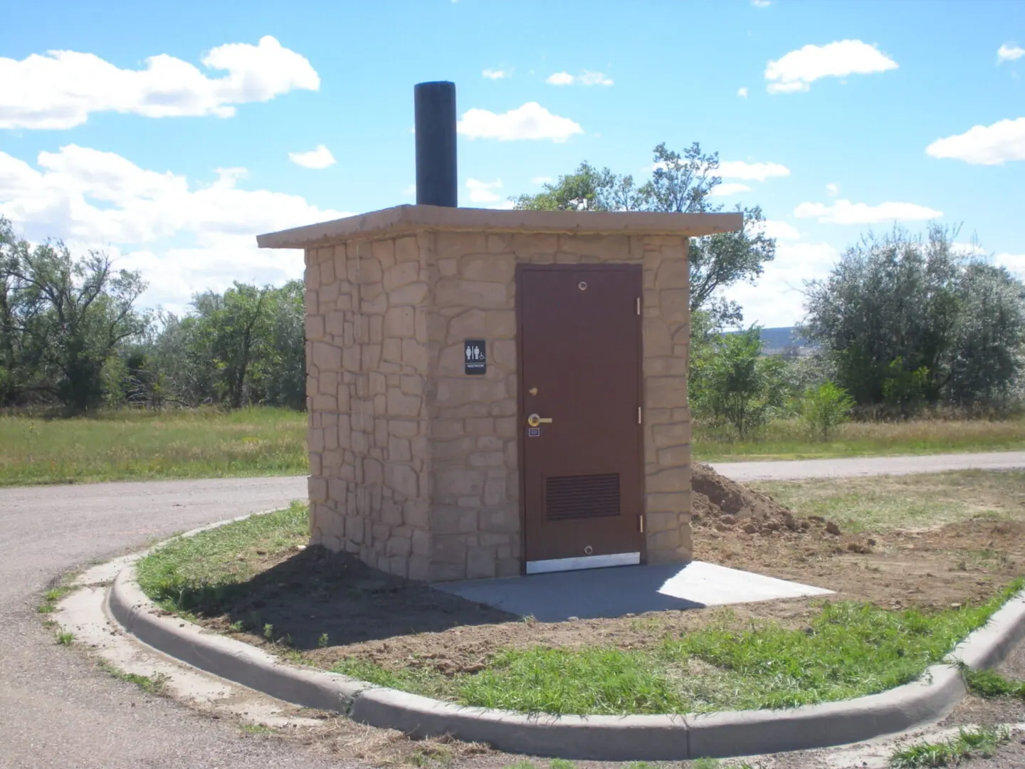 A small stone rest area bathroom with a brown door and a chimney, situated on a circular pavement, surrounded by grass and trees under a partly cloudy sky.