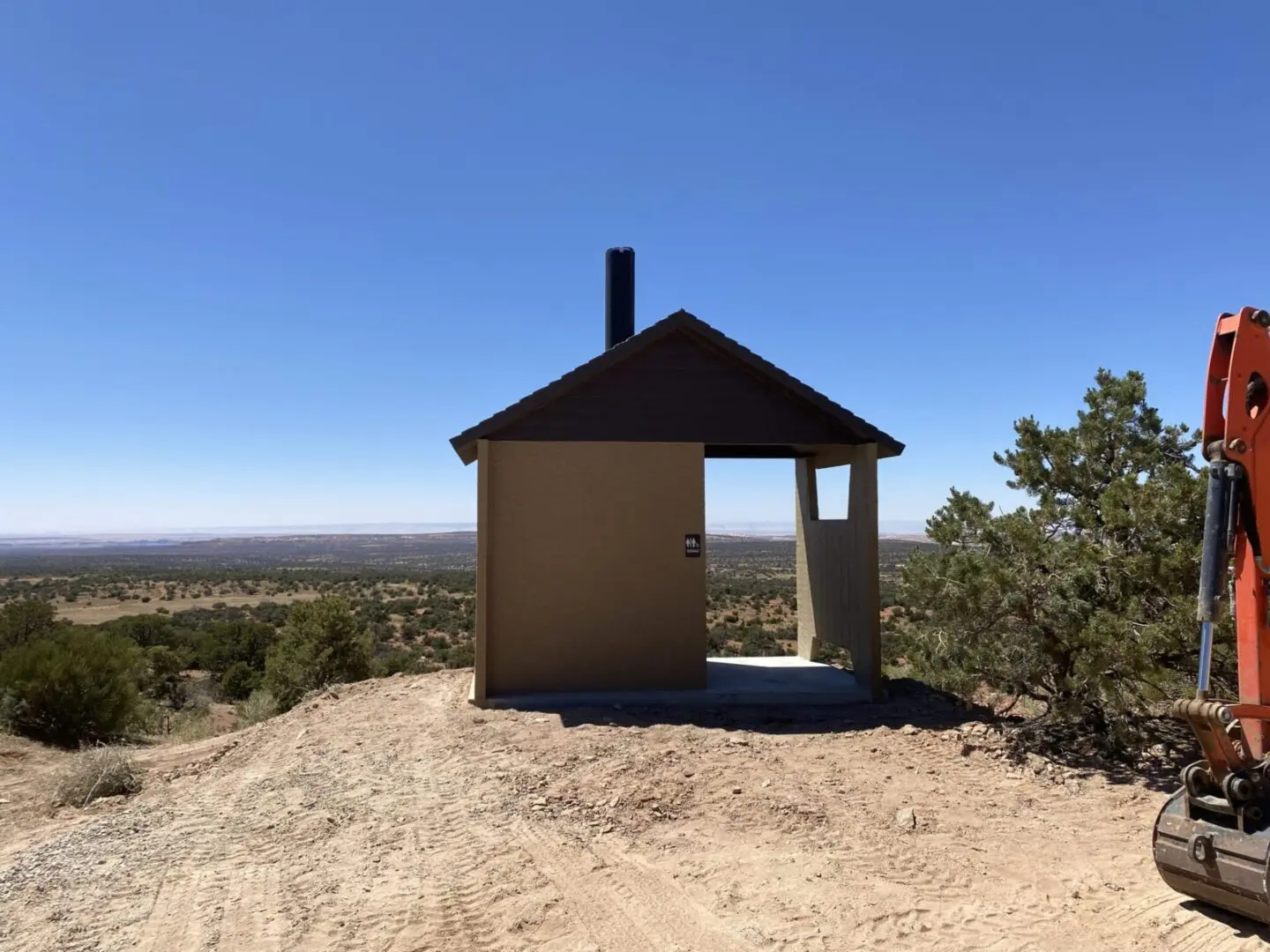 A small outhouse with a slanted roof stands on a dirt clearing in a rural area, surrounded by sparse vegetation under a clear blue sky.