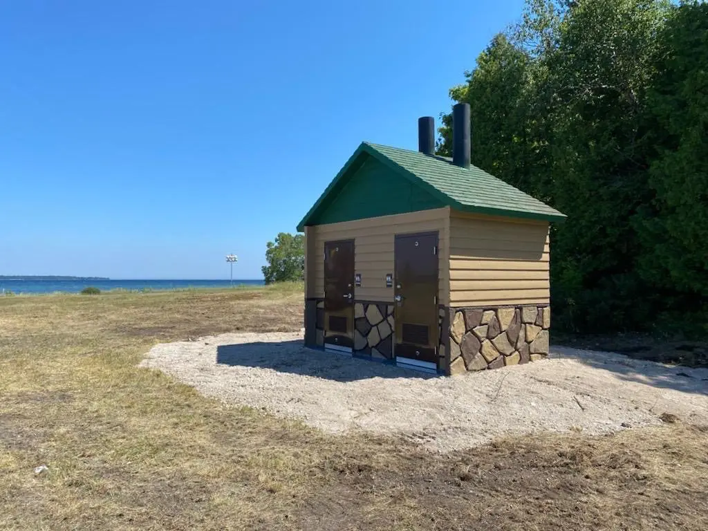 A small, rustic restroom building with green roof stands on a grassy area near a body of water, surrounded by trees.