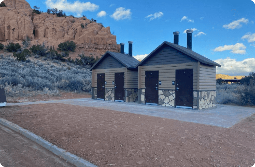 Two small restroom buildings with brown siding and black doors are set on a concrete slab in a desert landscape with rock formations and sparse vegetation under a blue sky.