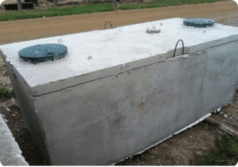 A large rectangular concrete septic tank with two circular green lids is positioned outdoors on gravel and grass.